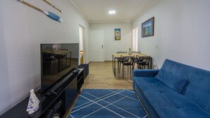 A living room with a TV, blue sofa, dining table, chairs, and a blue rug. - Apto a 750m da Praia da Enseada com Ar e Wi-fi (Guarujá)