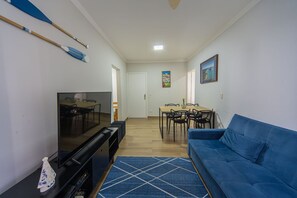 A living room with a TV, blue sofa, dining table, chairs, and a blue rug. - Apto a 750m da Praia da Enseada com Ar e Wi-fi (Guarujá)