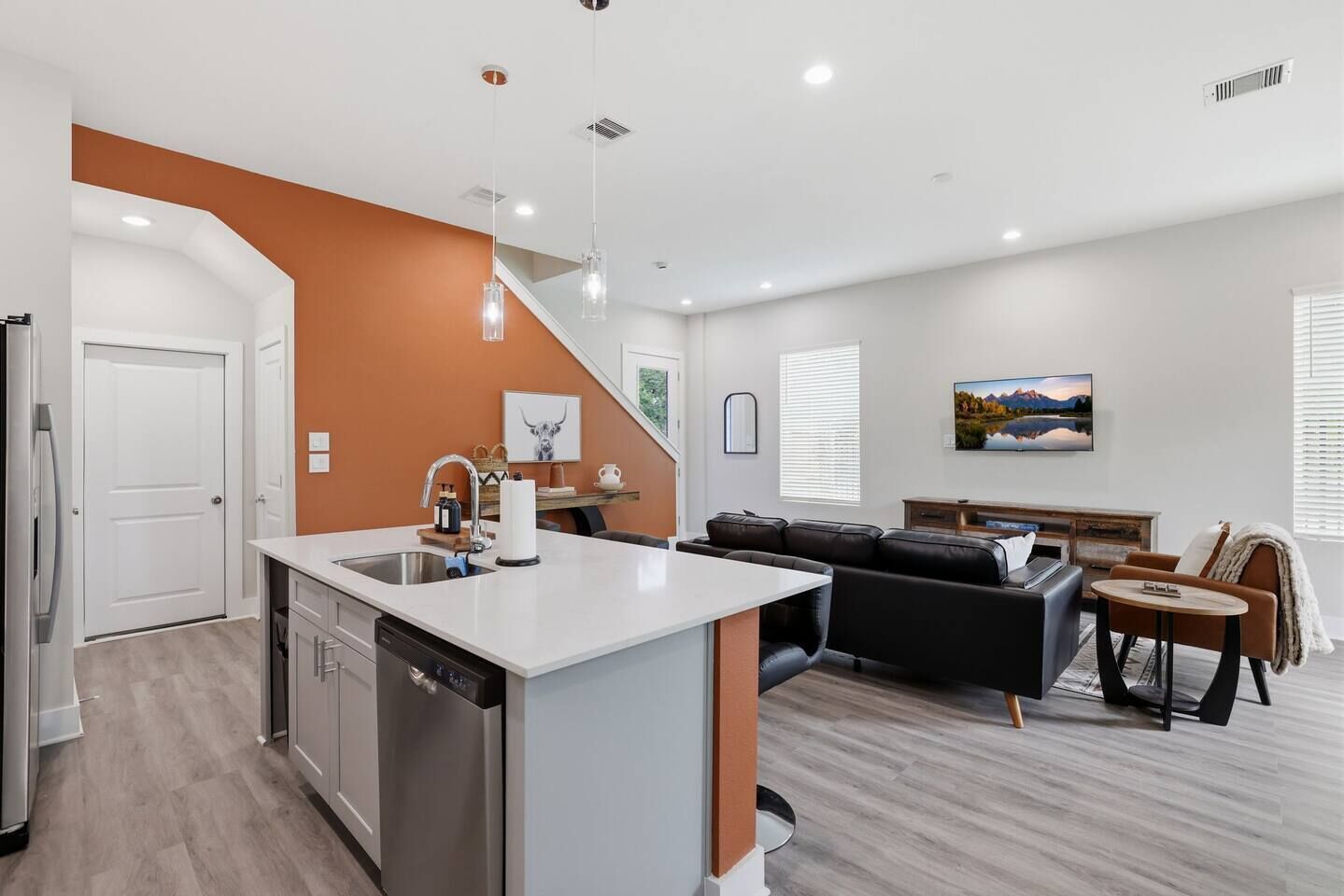 A modern apartment with a kitchen island, a dining area, a living room with a sofa and a TV, and a staircase.