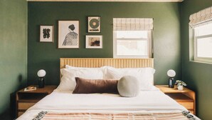 Ein Schlafzimmer mit einem Bett, zwei Nachttischen, einem Fenster mit Jalousien und drei gerahmten Kunstwerken an der Wand.