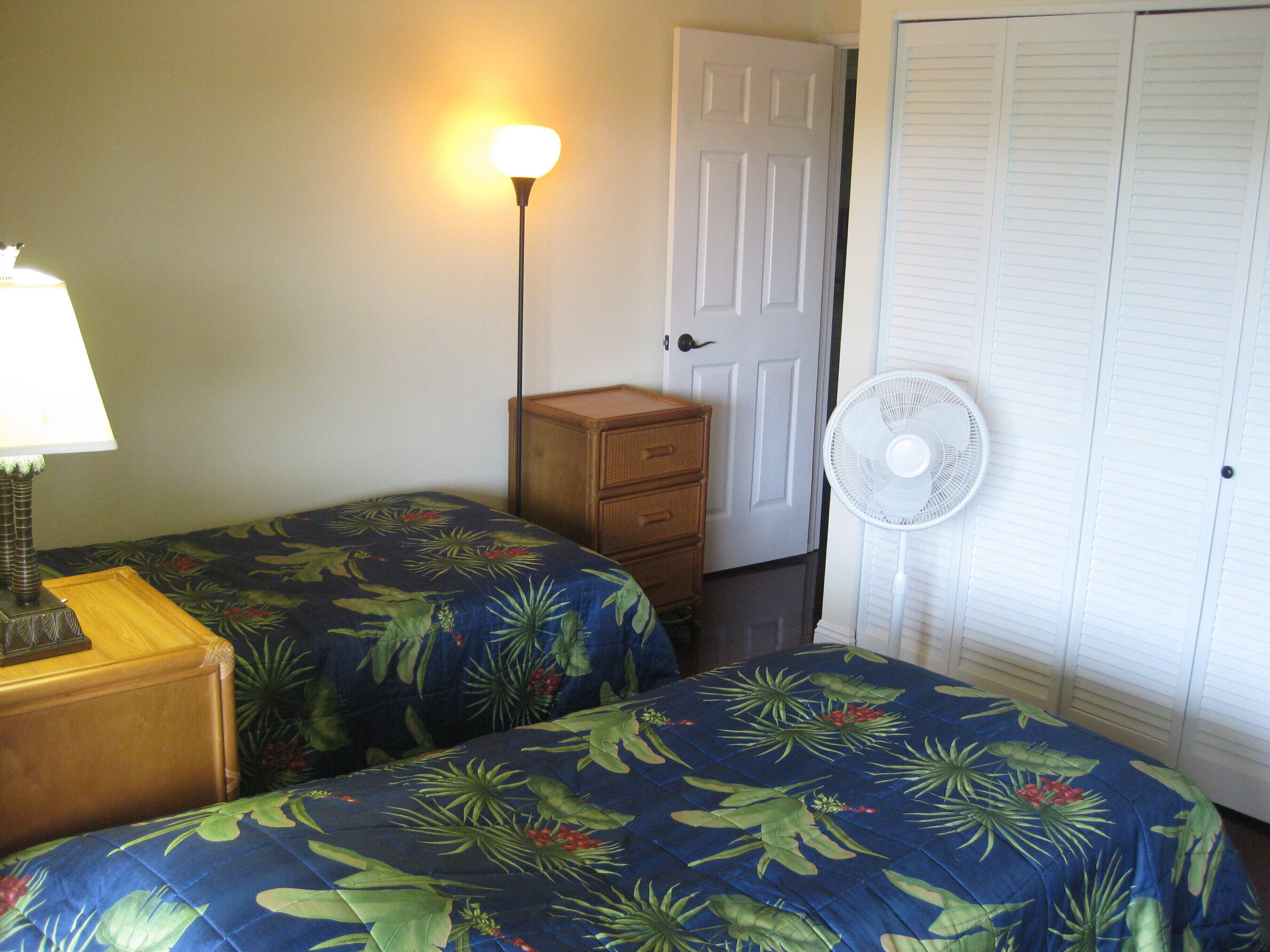 Une chambre avec deux lits, une table de chevet, une lampe, un ventilateur et une porte.