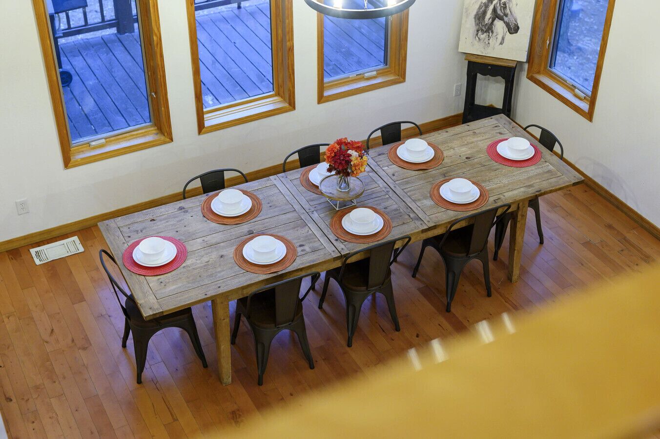 Une salle à manger avec une table en bois dressée pour six personnes, entourée de chaises, et de grandes fenêtres à cadres en bois.