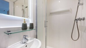 A bathroom with a glass shelf holding two bottles, a white sink, and a shower with a hose. - For Students Only Holden Court in Brighton (Brighton)