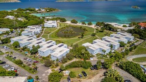 Vista aérea de uma área residencial costeira com vários edifícios de telhado azul, um parque circular central e uma vista clara do oceano e montanhas ao longe.