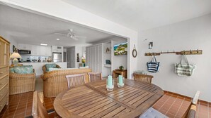 Uma área de refeições com uma mesa redonda de madeira, móveis de vime e uma cozinha ao fundo.