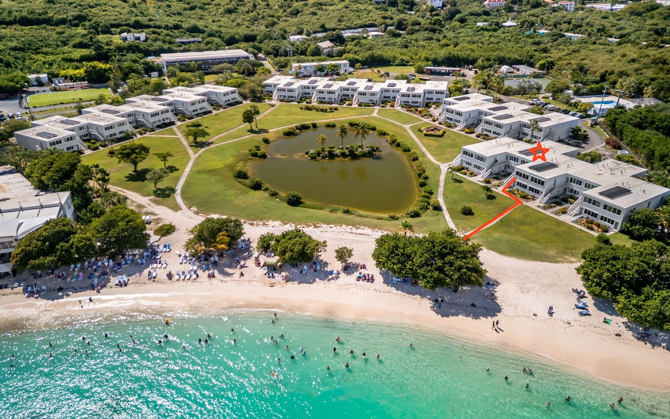 An aerial view of a resort with multiple buildings, a central pond, and a beach area.