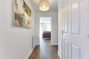 A hallway with a radiator, a painting on the wall, and a door leading to a room with a bed. - The Fox Den - Chelmsford | 3-bed Stay (Broomfield)