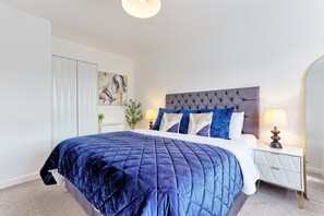A bedroom with a bed, a nightstand, a lamp, a mirror, and a painting on the wall. - The Fox Den - Chelmsford | 3-bed Stay (Broomfield)