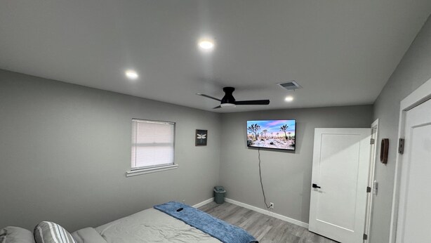 Ein Schlafzimmer mit einem Bett, einem Deckenventilator, einem an der Wand montierten Fernseher, einem Fenster mit Jalousien und einer Tür.