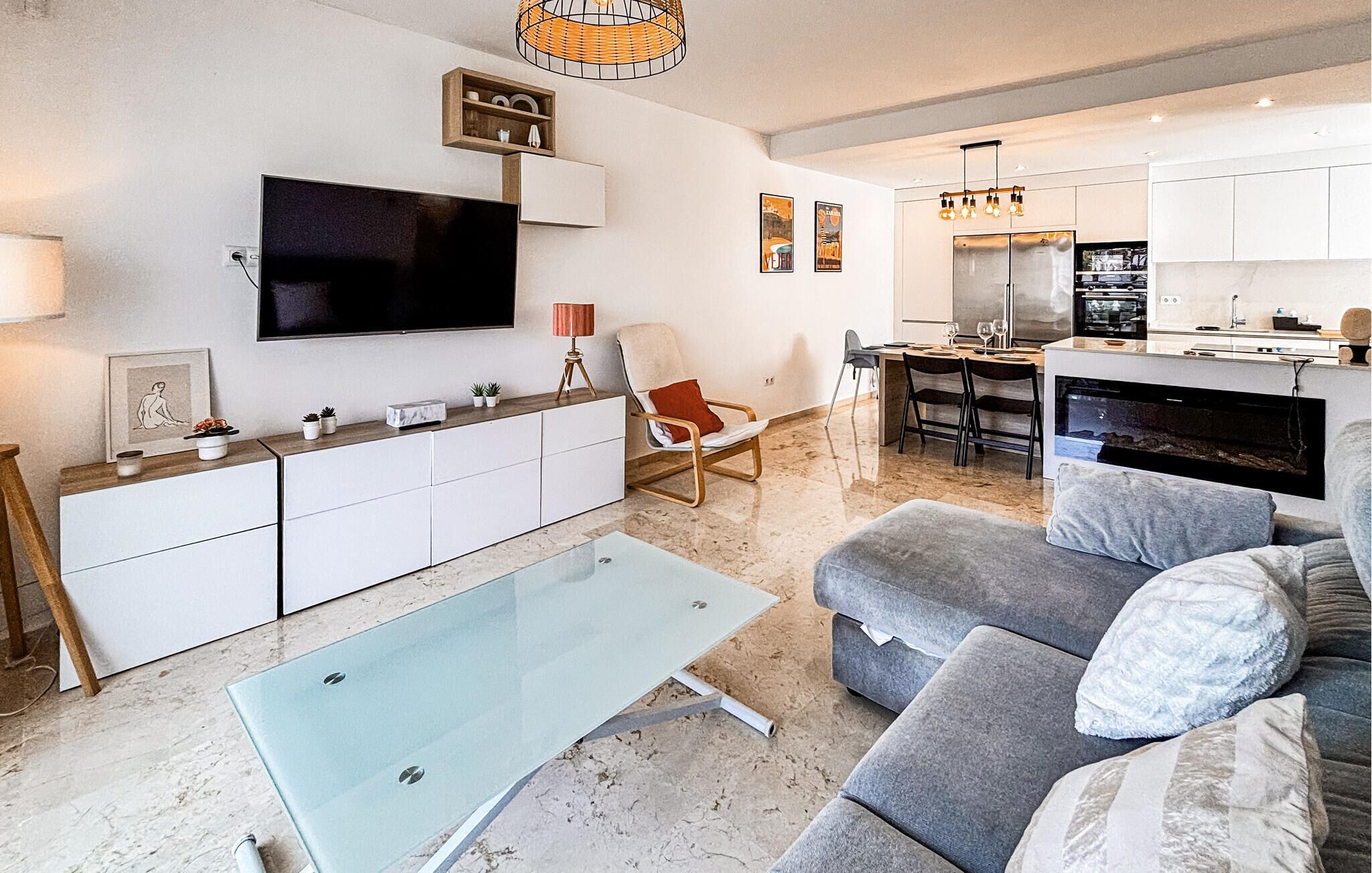 A modern living room with a glass coffee table, grey sofa, and a flat-screen TV mounted on the wall.