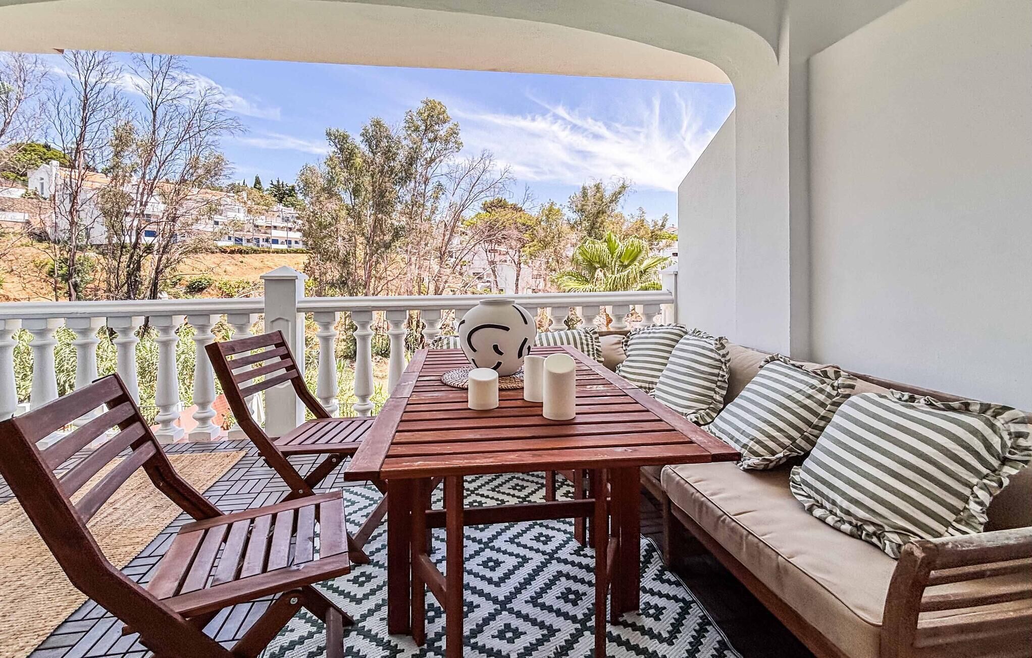 A balcony with a wooden table and chairs, a sofa with striped cushions, and a decorative vase.
