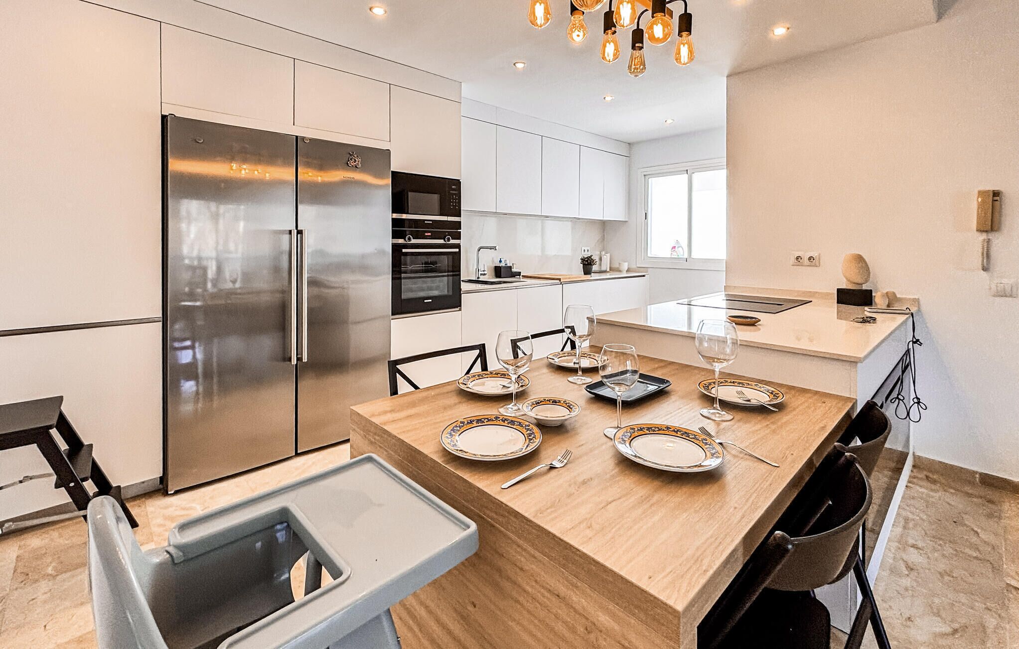 Une cuisine moderne dotée d’appareils électroménagers en acier inoxydable, une table à manger dressée avec des assiettes et des verres, et un luminaire avec des ampoules apparentes.