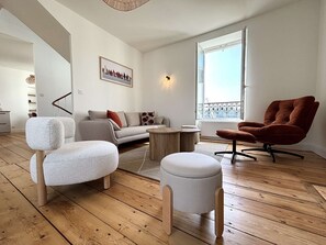 Uma sala de estar moderna com sofá, banqueta e poltrona, piso de madeira, janela com vista e um quadro na parede.