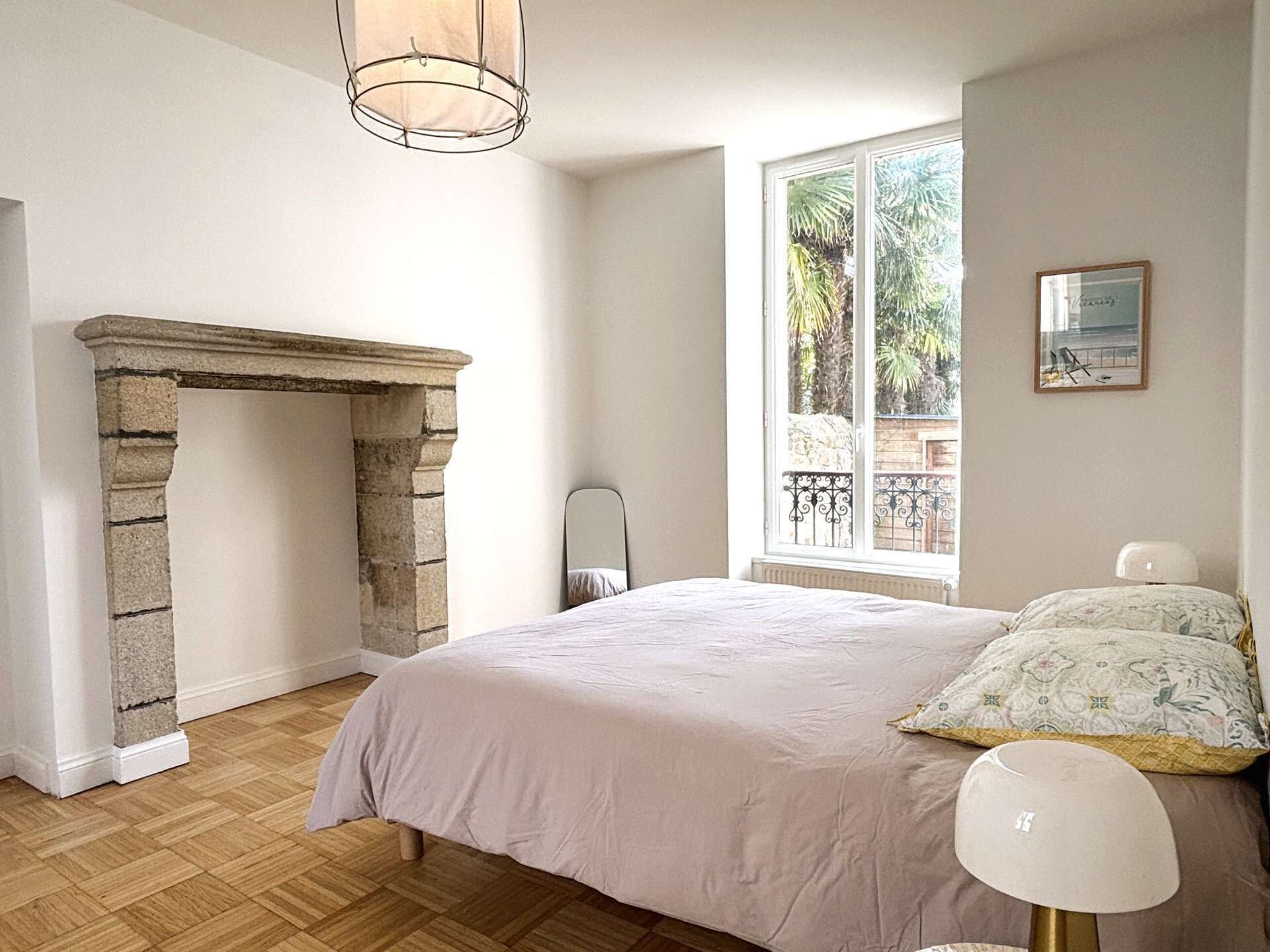 A bedroom with a stone fireplace, a bed with a white bedspread, a window with a view of palm trees, and a framed picture on the wall.