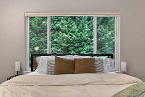 Ein Schlafzimmer mit großem Fenster, einem Bett mit weißem und gemustertem Kopfkissen und Nachttischlampen.
