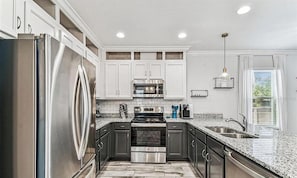 Una cocina moderna con electrodomésticos de acero inoxidable, armarios blancos, encimeras de granito y una ventana con persianas.