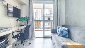 Un apartamento moderno con televisor de pantalla plana, un escritorio con computadora, un sofá gris con cojines azules y un balcón con vistas a edificios altos.