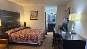 Habitación de hotel con cama, escritorio, televisor, lámpara, espejo y kitchenette.