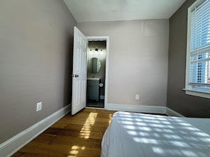 A bedroom with a bed, a window with blinds, a door leading to a bathroom, and a light switch on the wall.