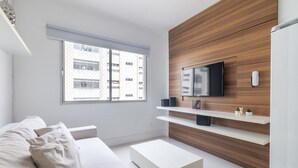 Ein modernes Wohnzimmer mit einer Couch, einem Couchtisch, einem an einer Holzwand montierten Fernseher und einem Fenster mit Blick auf ein Gebäude.