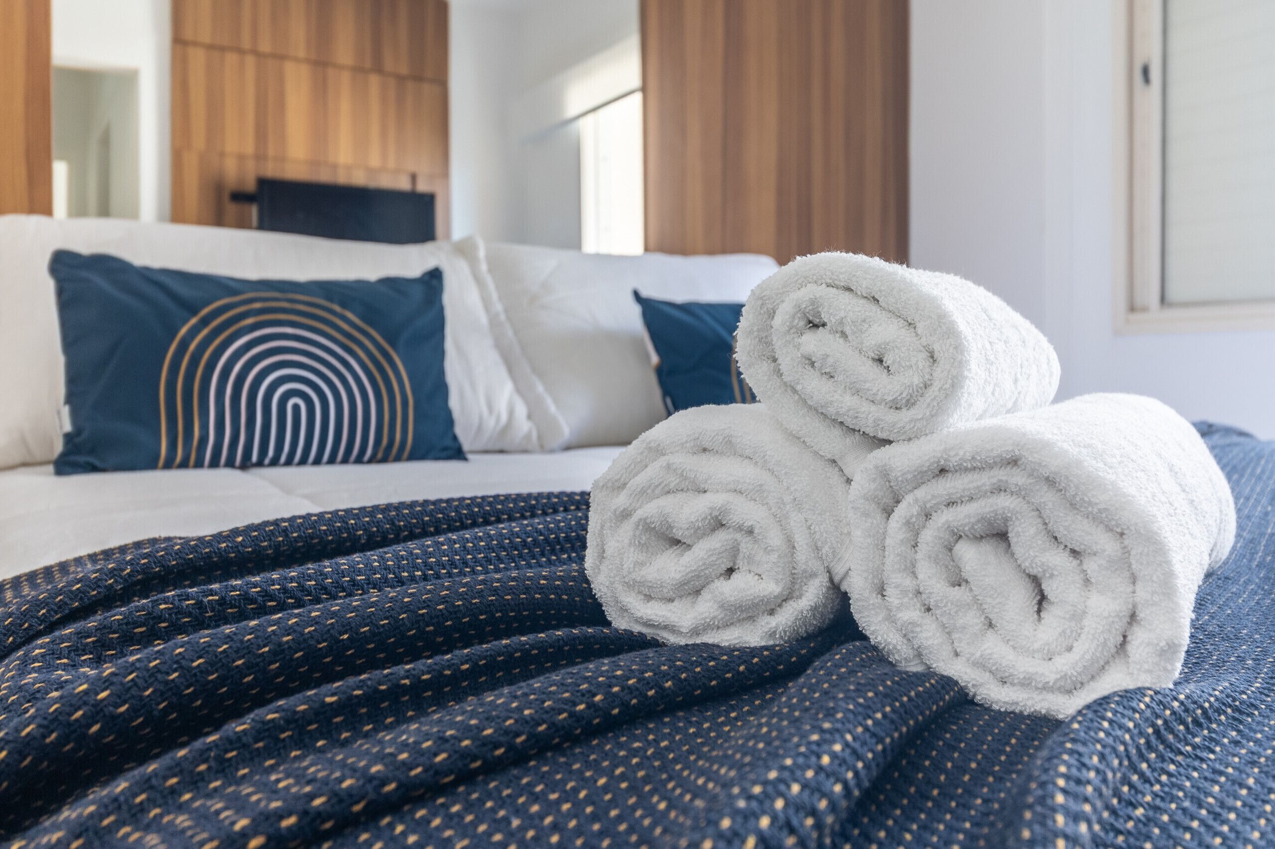 A neatly made bed with rolled white towels, a dark blue patterned blanket, and a blue pillow with a concentric circle design.