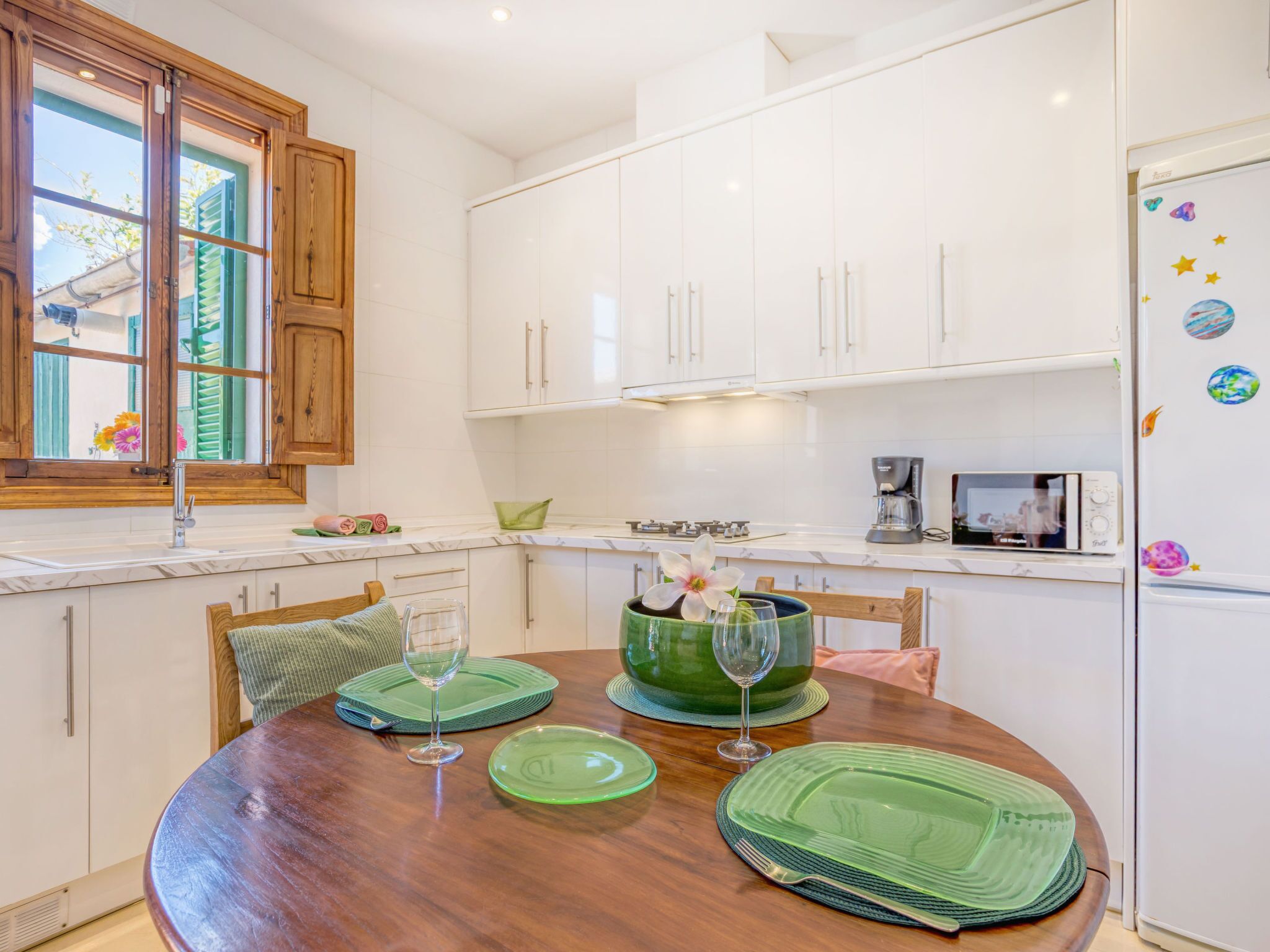 Una cocina moderna con armarios blancos, una mesa de comedor de madera con individuales verdes y un refrigerador con imanes decorativos.