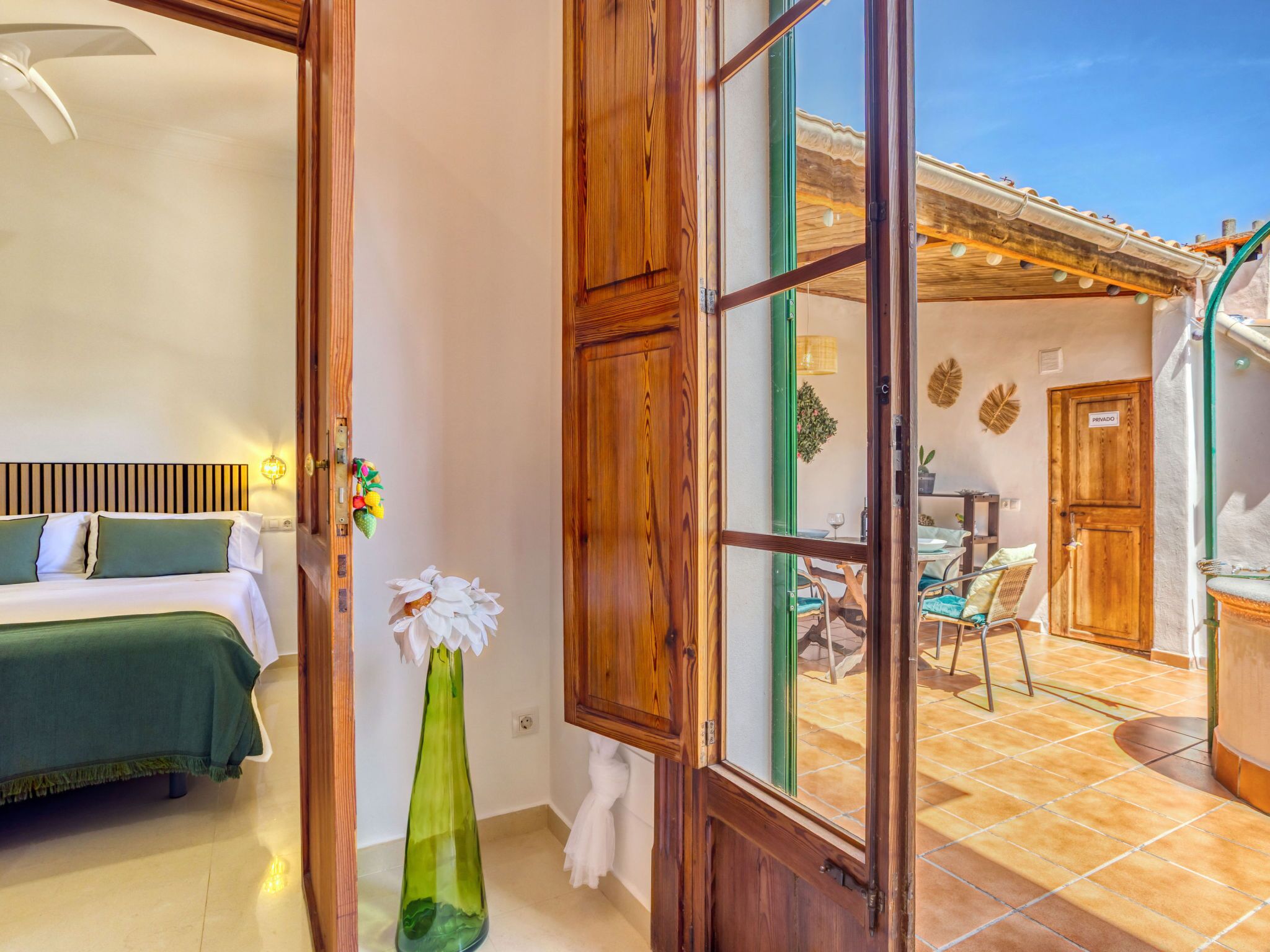Una stanza con un letto, un vaso verde con un fiore bianco e una porta in legno che dà su un'area esterna con un tavolo e delle sedie.