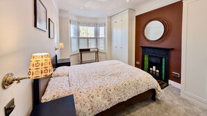 A bedroom with a bed, a nightstand, a lamp, a fireplace, and a window with shutters. - Cosy Bright 2-bed House in Earlsfield (London)