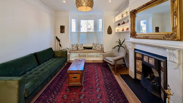 A living room with a green sofa, a wooden coffee table, a patterned rug, a fireplace, and a large ornate mirror. - Cosy Bright 2-bed House in Earlsfield (London)