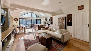 A spacious living area with a large sofa, a coffee table, and a TV. There is a kitchen area with cabinets and a refrigerator. The room has a high ceiling with skylights and a large window providing natural light. - Cosy Bright 2-bed House in Earlsfield (London)