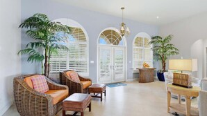 Un salón amplio con muebles de mimbre, un candelabro y ventanas grandes con contraventanas.