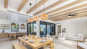 Eine moderne Küche mit Holzbalken, einem Essbereich mit einem Holztisch, gedeckt für sechs Personen, und einem weißen Sofa in einem Wohnbereich mit großen Fenstern.