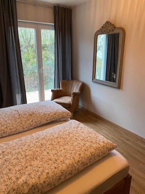 A room with two beds, a chair, a mirror, and a view of greenery outside. - Apartment 'Rebblick' with Mountain View, Balcony and Wi-Fi (Müllheim im Markgräflerland)