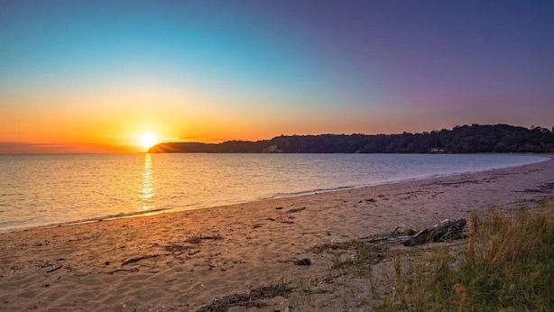 Ein Strand bei Sonnenuntergang, ruhiges Wasser und eine entfernte Baumlinie.