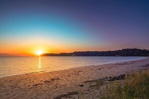Une plage au coucher du soleil, avec une eau calme et une ligne d’arbres à l’horizon.