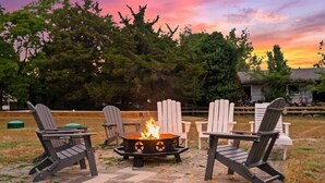 Ein Feuerkorb mit vier Adirondack-Stühlen, die darum angeordnet sind, auf einer Steinterrasse, im Hintergrund Bäume und ein Holzzaun.