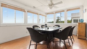 Ein Zimmer mit einem runden Tisch, Korbstühlen, einem Deckenventilator und großen Fenstern.