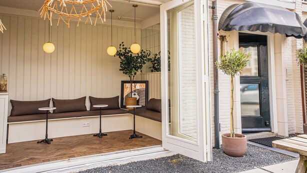 Un coin salon accueillant avec un banc et des tables, un lustre en osier et une plante en pot devant un bâtiment sur lequel est apposée une enseigne.