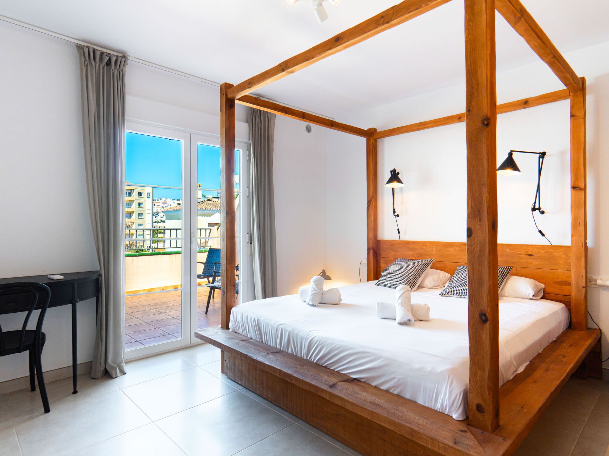 A four-poster bed with a wooden frame, white bedding, and a view of a cityscape through a sliding glass door.