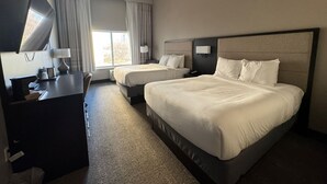 Habitación de hotel con una cama grande, una cama más pequeña, un escritorio, un televisor y una ventana con cortinas.