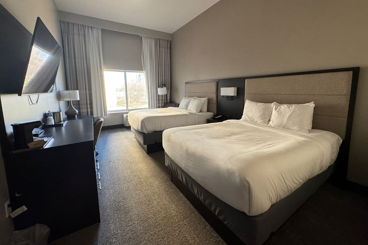 Ein Hotelzimmer mit einem großen Bett, einem kleineren Bett, einem Schreibtisch, einem Fernseher und einem Fenster mit Vorhängen.