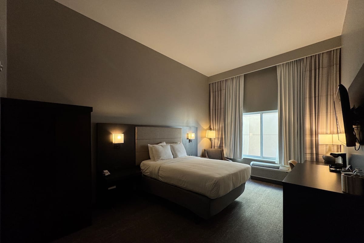 Ein Hotelzimmer mit Bett, Nachttischlampen, einem Schreibtisch und einem großen Fenster mit Vorhängen.