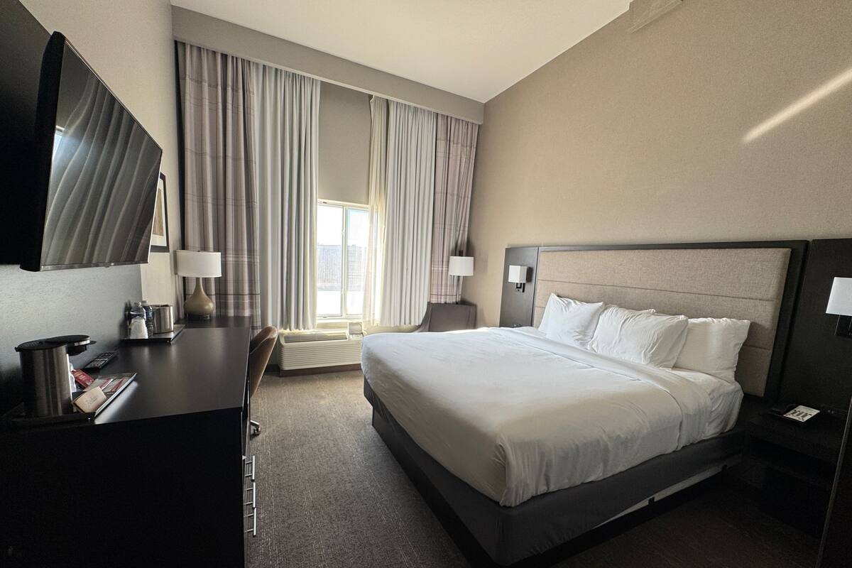 Ein Hotelzimmer mit einem großen Bett, einem Schreibtisch, einem Sessel, einem Fernseher und einem Fenster mit Vorhängen.