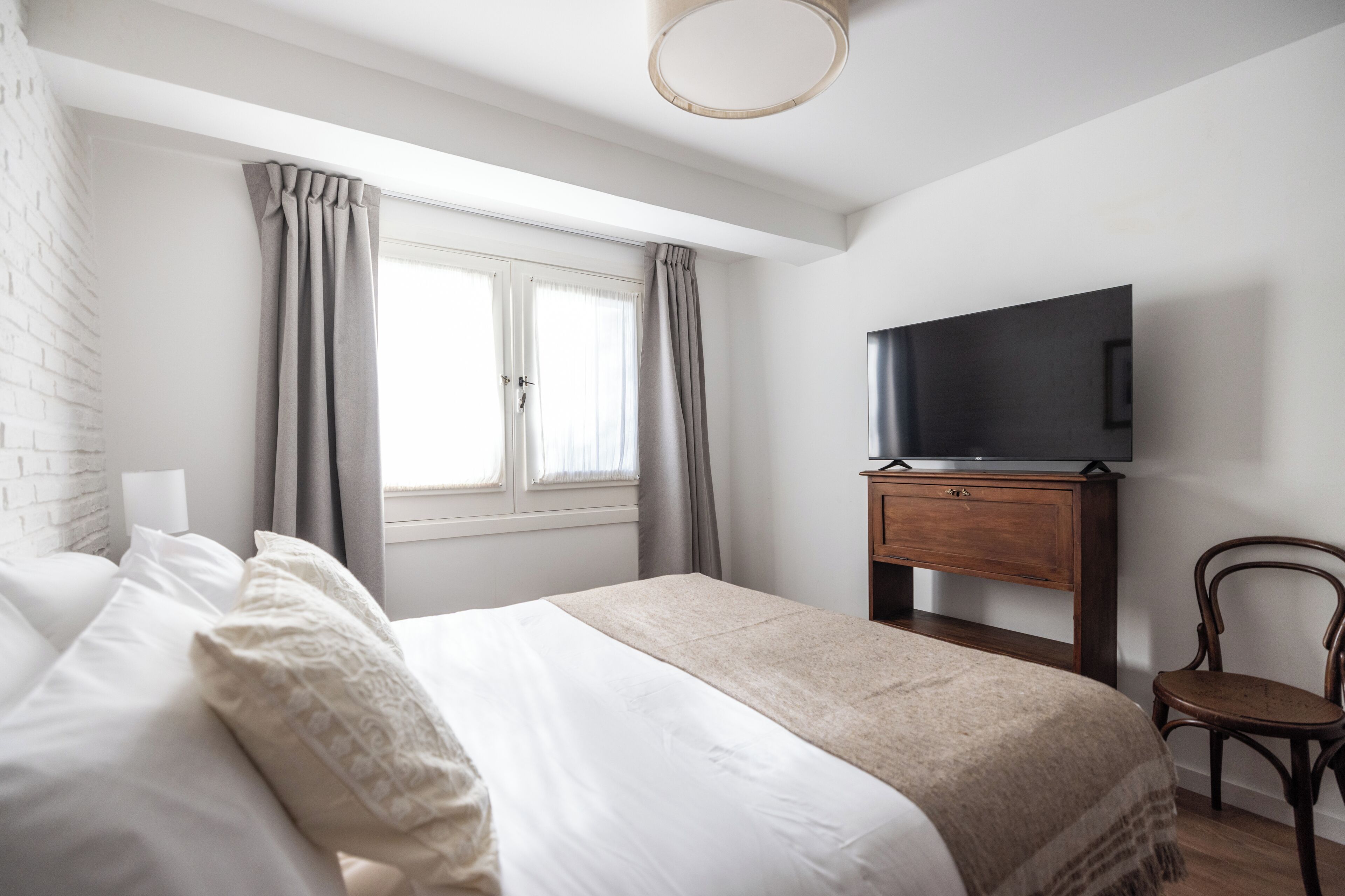 A bedroom with a bed, a TV on a wooden stand, a chair, and a window with curtains.