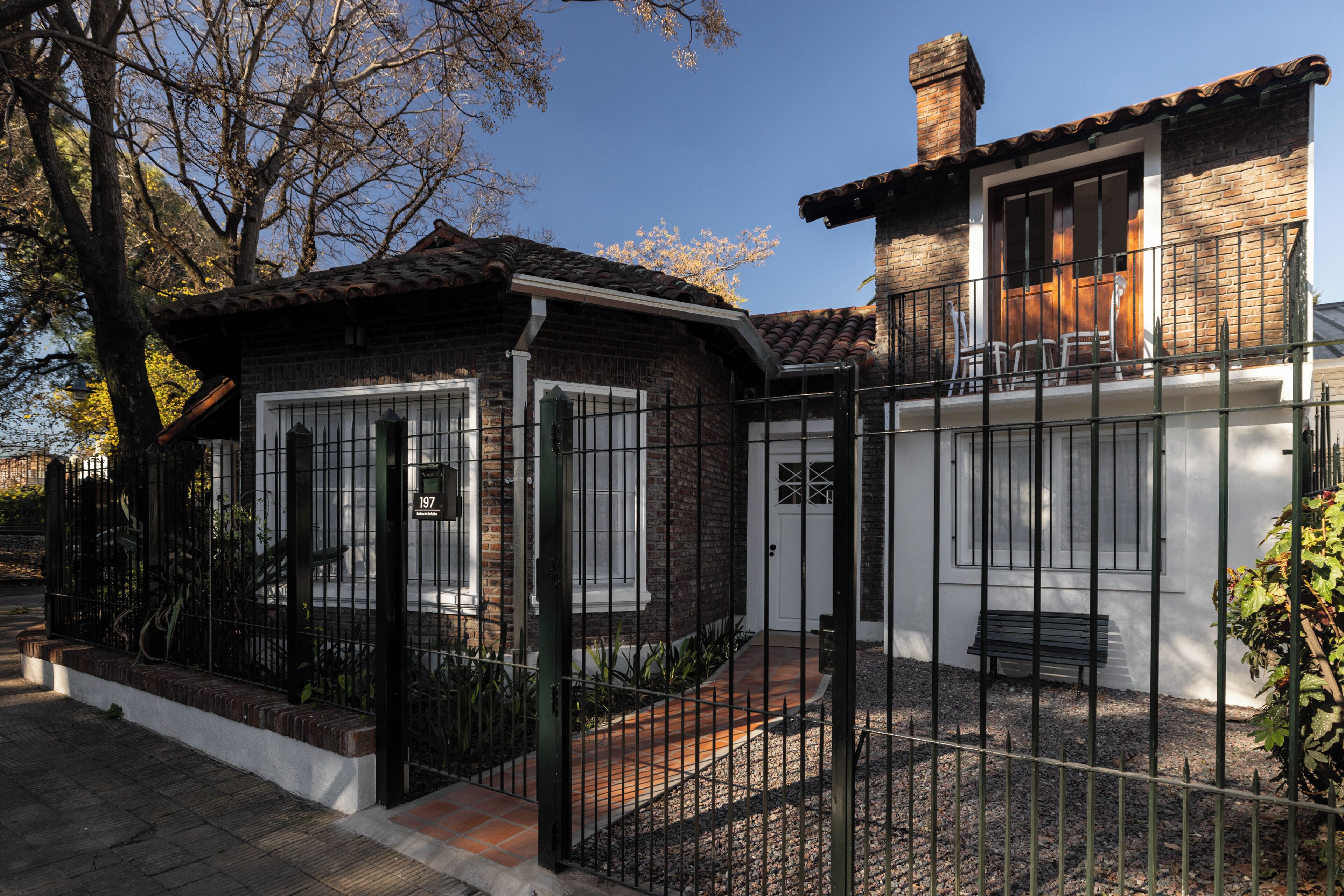 Une maison à deux étages, avec une façade en briques, une clôture en métal noir et un petit porche doté d’un banc.