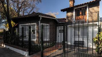 Une maison à deux étages, avec une façade en briques, une clôture en métal noir et un petit porche doté d’un banc.