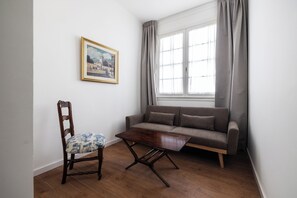 Habitació amb terra de fusta, un sofà, una cadira, una taula, una finestra amb cortines i un quadre al mur.