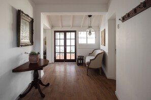 A hallway with a wooden floor, a white wall with a framed picture, a round table with a potted plant, a white armchair, and a door leading to an outdoor area. - Acassuso Suites (Acassuso)
