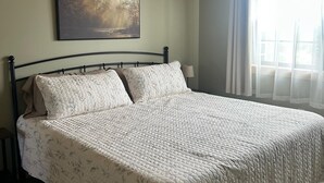 Une chambre avec un plafond en bois, un lit recouvert d’une courtepointe matelassée, un tableau encadré au mur et une fenêtre ornée de rideaux légers.