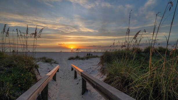 Un sentiero di legno conduce a una spiaggia con un tramonto sullo sfondo.